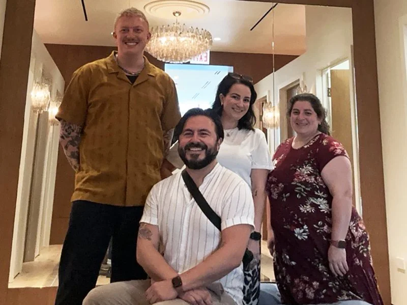 Four people posing together indoors, smiling, with a chandelier hanging from the ceiling in the background.