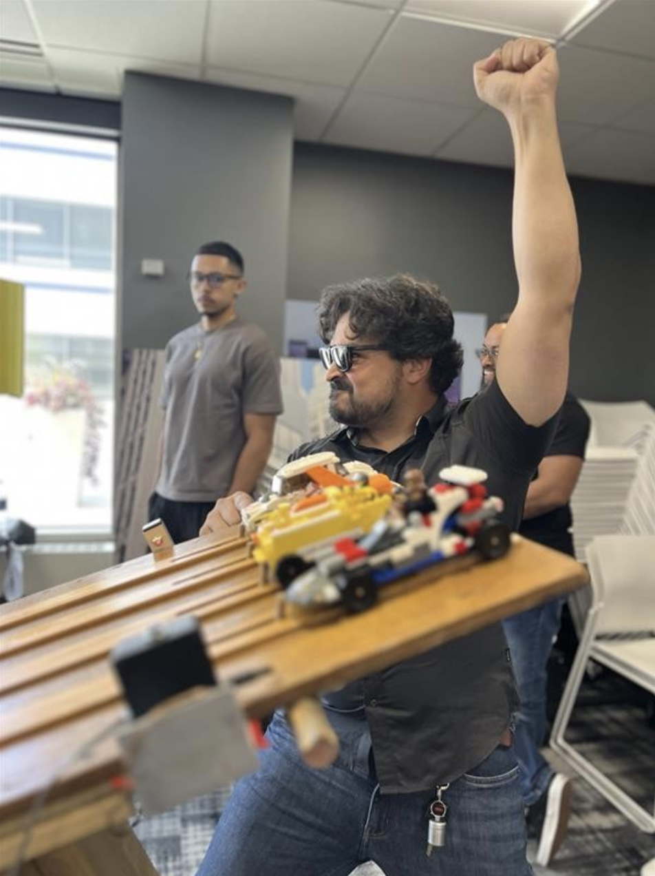 Man with dark, curly hair and glasses raising his right fist in triumph at a LEGO car racing event, with LEGO cars on a wooden track in the foreground and several onlookers in the background.