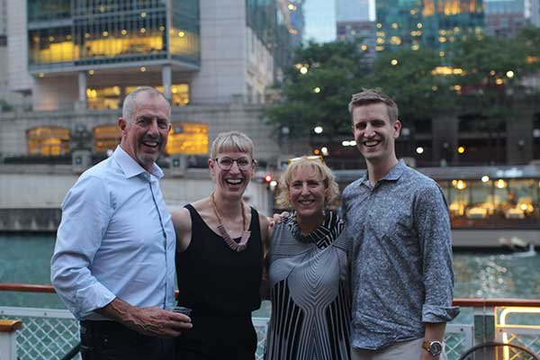 Four people standing together outdoors with city buildings and water in the background, smiling for the camera.