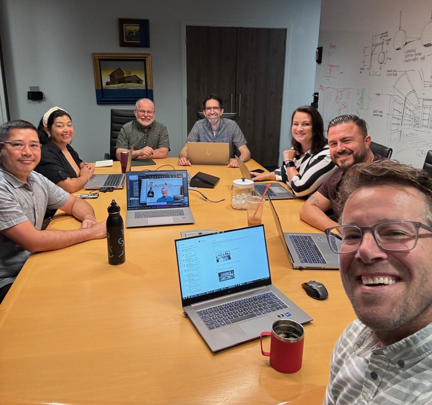 A group of eight diverse people sitting around a conference table in a meeting room, smiling for a group selfie, with laptops, a water bottle, and drinks on the table, and artwork and whiteboard drawings on the walls.