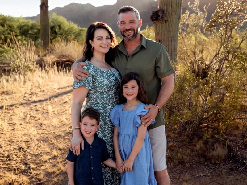 A smiling family of four standing outdoors with desert landscape, cacti, and mountains in the background during sunset, including a woman, a man, a young girl, and a young boy.