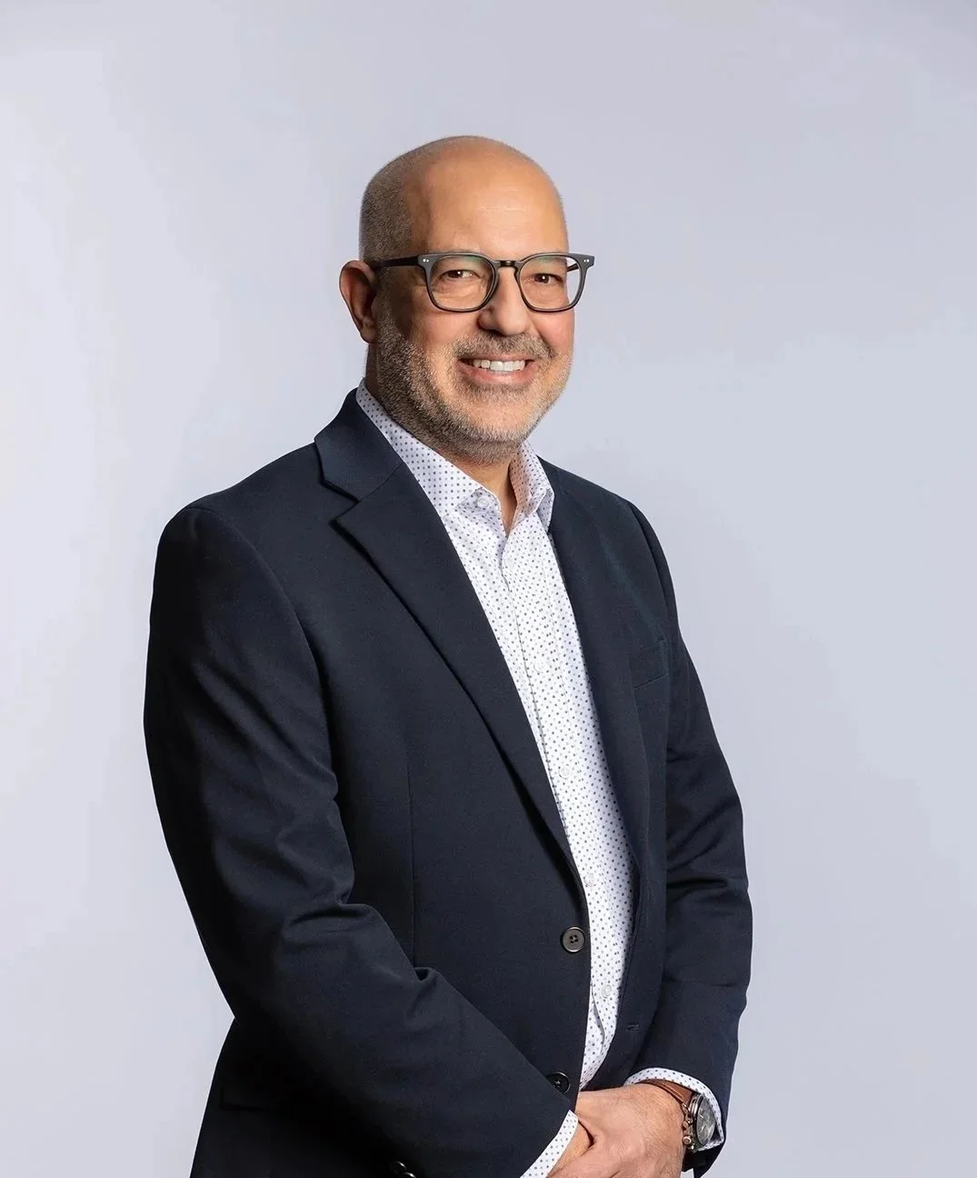 A man with glasses, a suit jacket, and a white polka dot shirt smiling and standing against a plain light gray background.