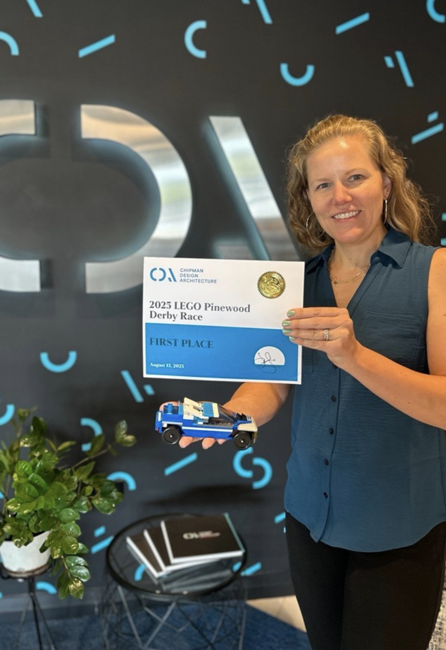 Woman holding a Lego car, a certificate for first place in the 2025 LEGO Pinewood Derby Race, and an award medal, smiling in front of a black background with blue clock graphics.