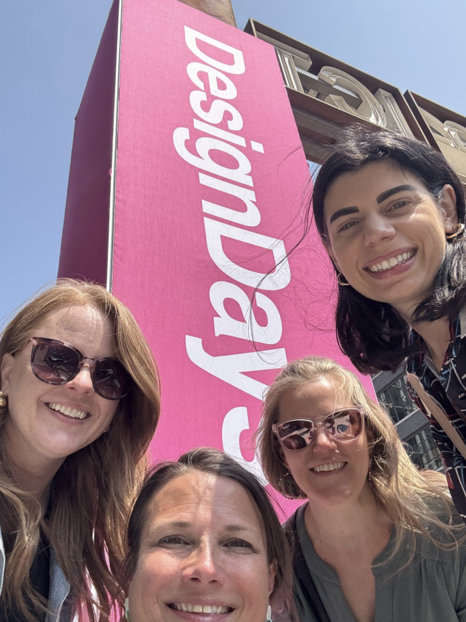 A group of five women taking a selfie in front of a large pink sign with white text that reads 'Advocates!' on a city street.