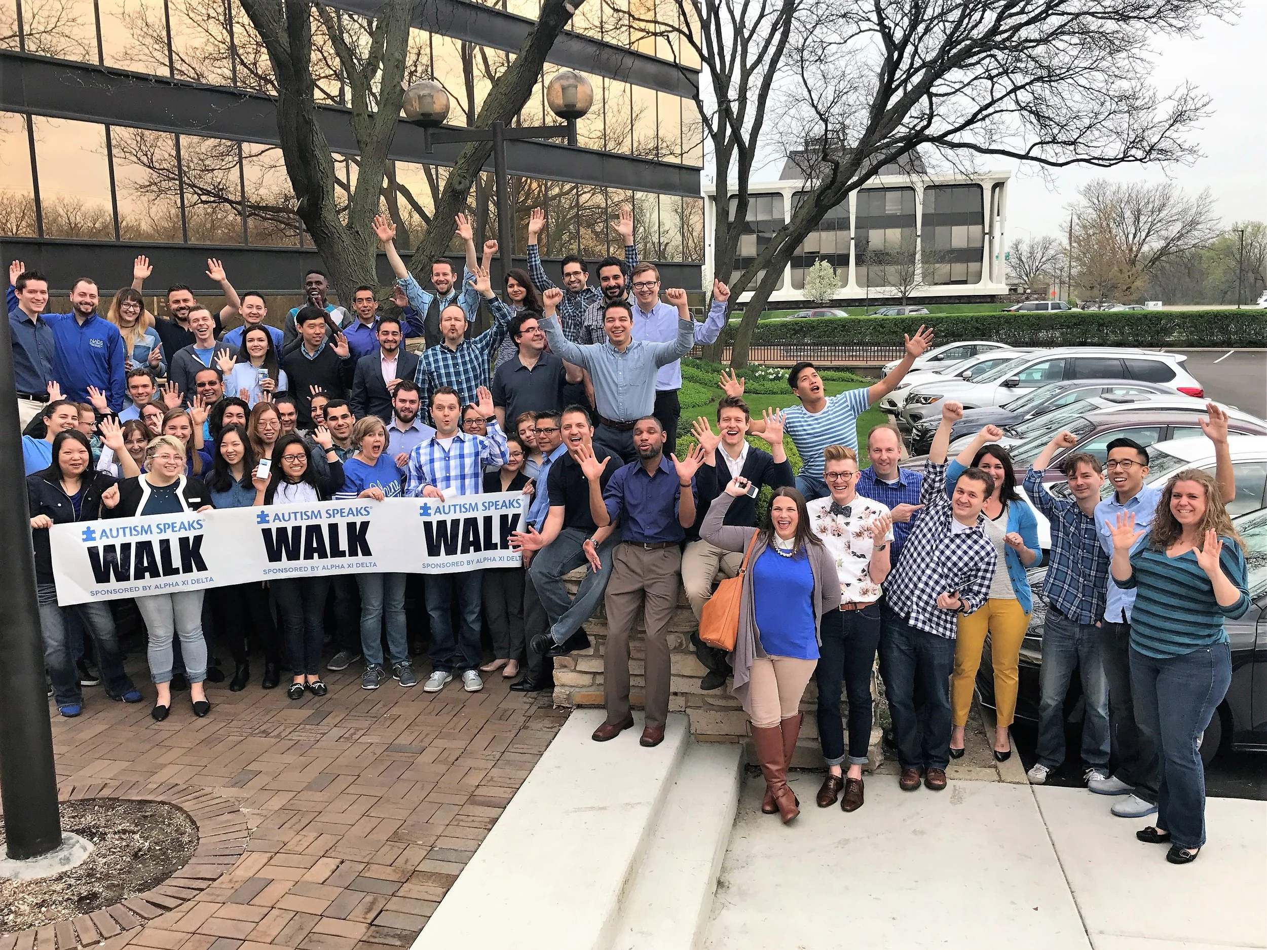 A large group of people gathered outdoors for an Autism speaks walk event, holding a banner, smiling, and raising their hands in celebration.