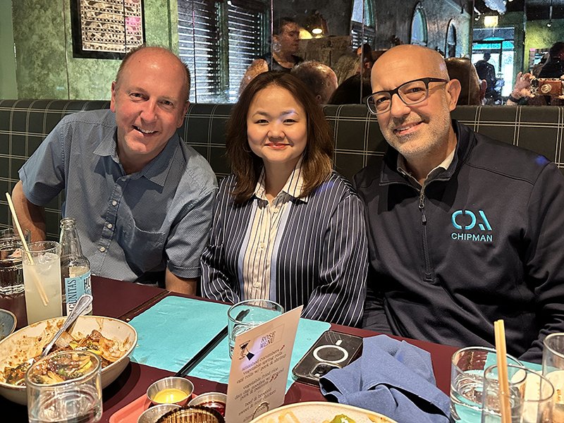 Three people sitting at a restaurant table smiling for the camera with food and drinks on the table.
