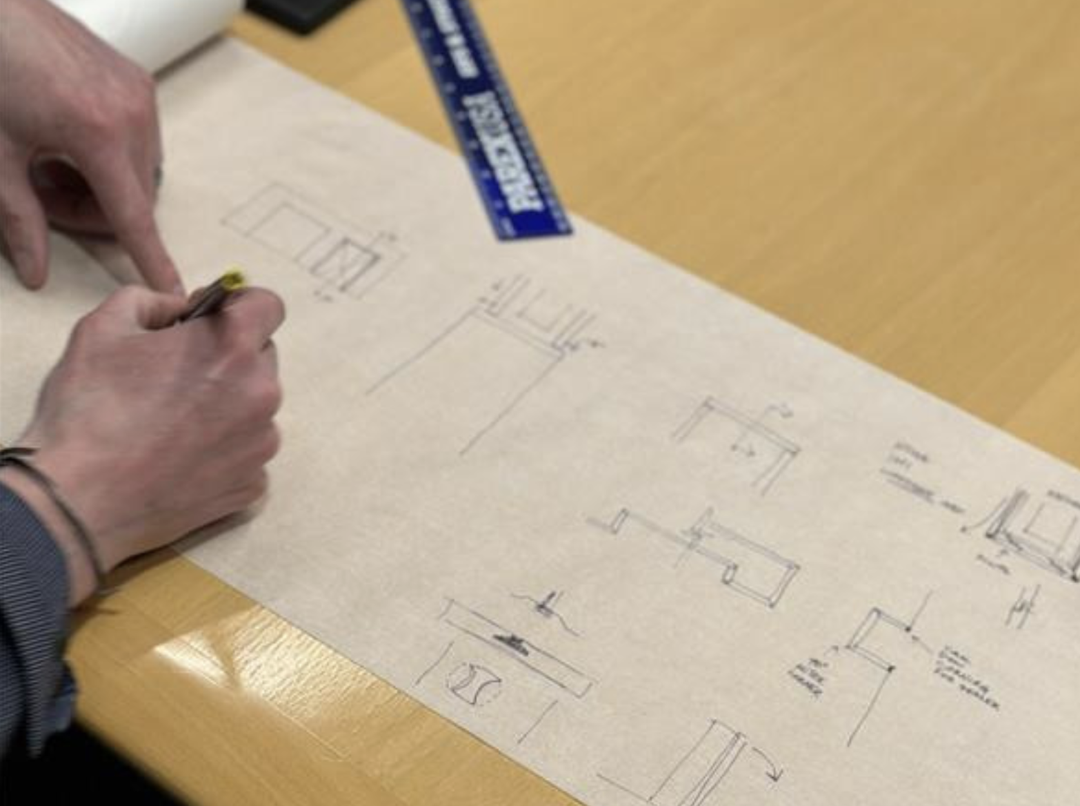 Person drawing architectural sketches and plans on paper with various room and furniture layouts, on a wooden table.