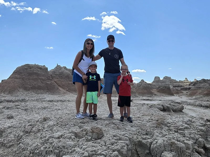 A family of five standing on a rocky landscape with layered rock formations in the background under a bright blue sky.