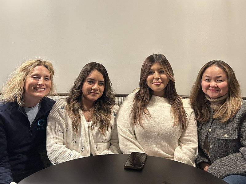 Four women sitting together at a table, smiling, with a phone on the table in front of them, against a plain light-colored wall.