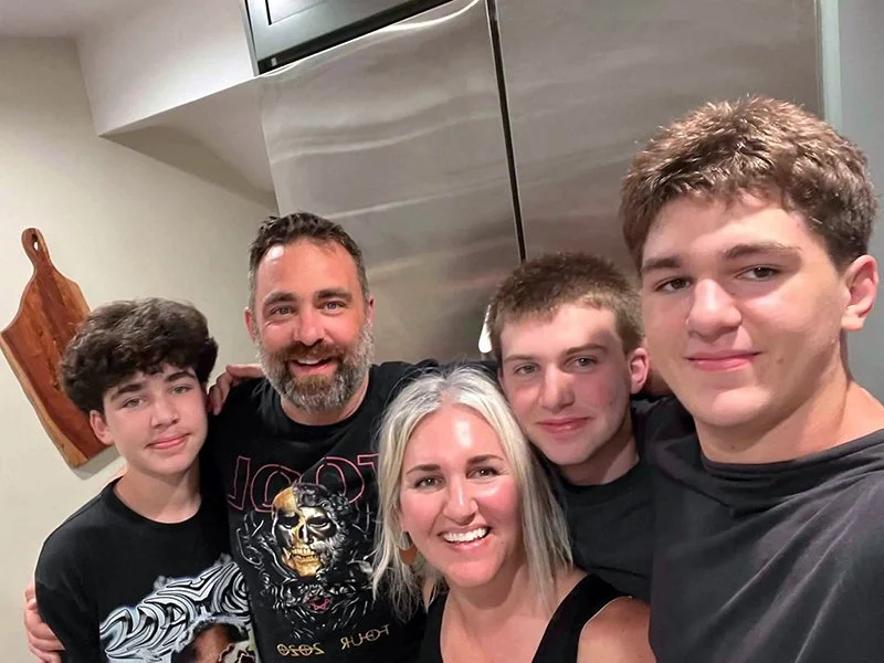 A family of five taking a selfie together in a kitchen. The family includes two adult men, one woman, and two teenage boys, all smiling at the camera.