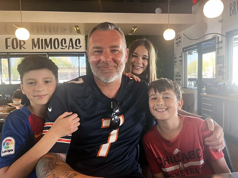 A man with a beard wearing a football jersey is sitting with three children in a restaurant, smiling at the camera.