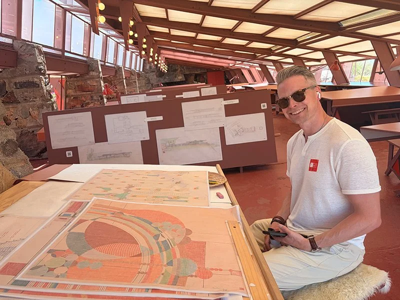 A man wearing sunglasses and a white t-shirt sitting at a table with architectural drawings and colorful artwork. He is smiling and holding a mobile device in one hand. The room has large windows, a slanted roof with exposed beams, and walls adorned with blueprints and sketches.