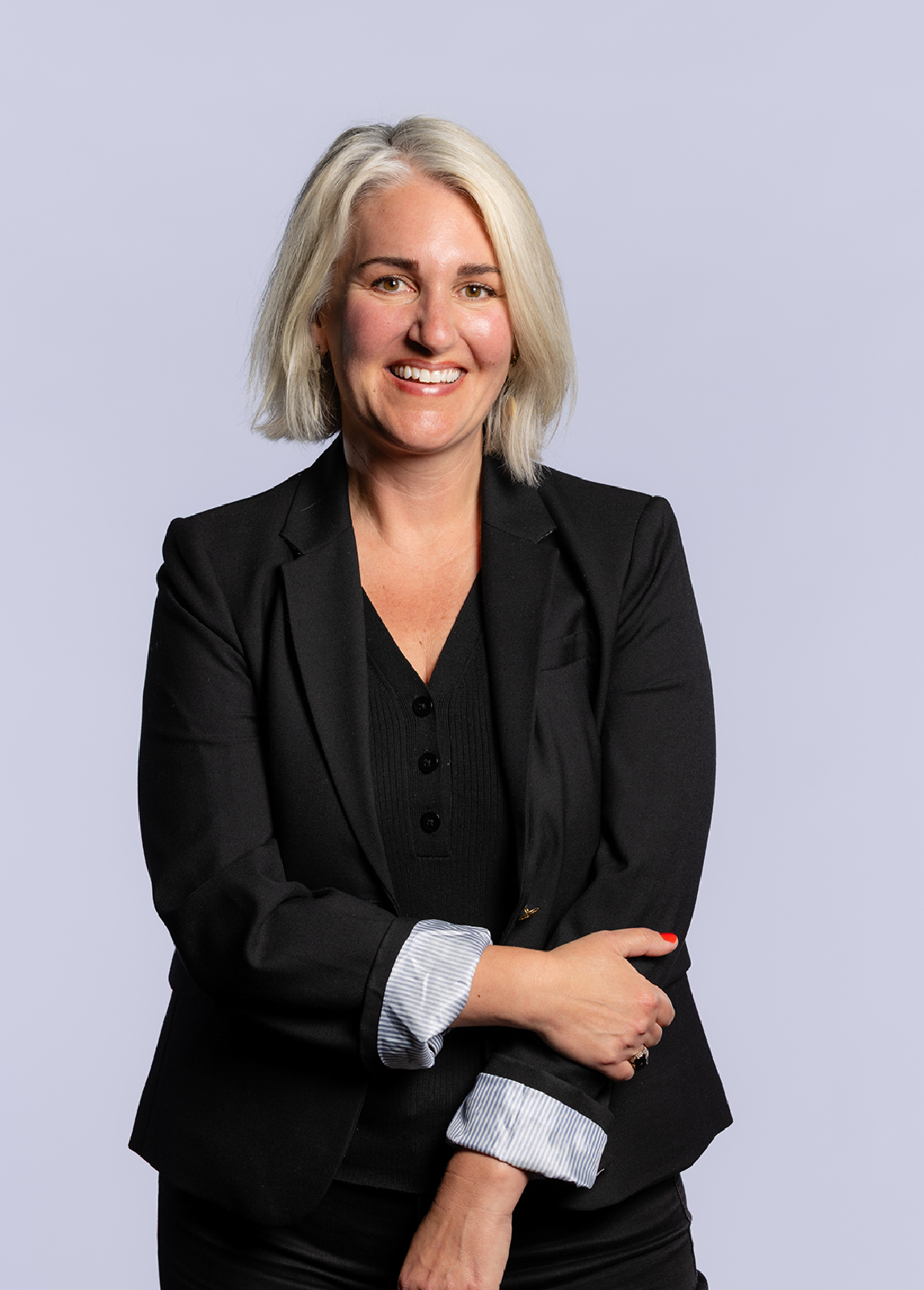 A woman with short blonde hair in a black blazer and black top smiling at the camera, against a light gray background.