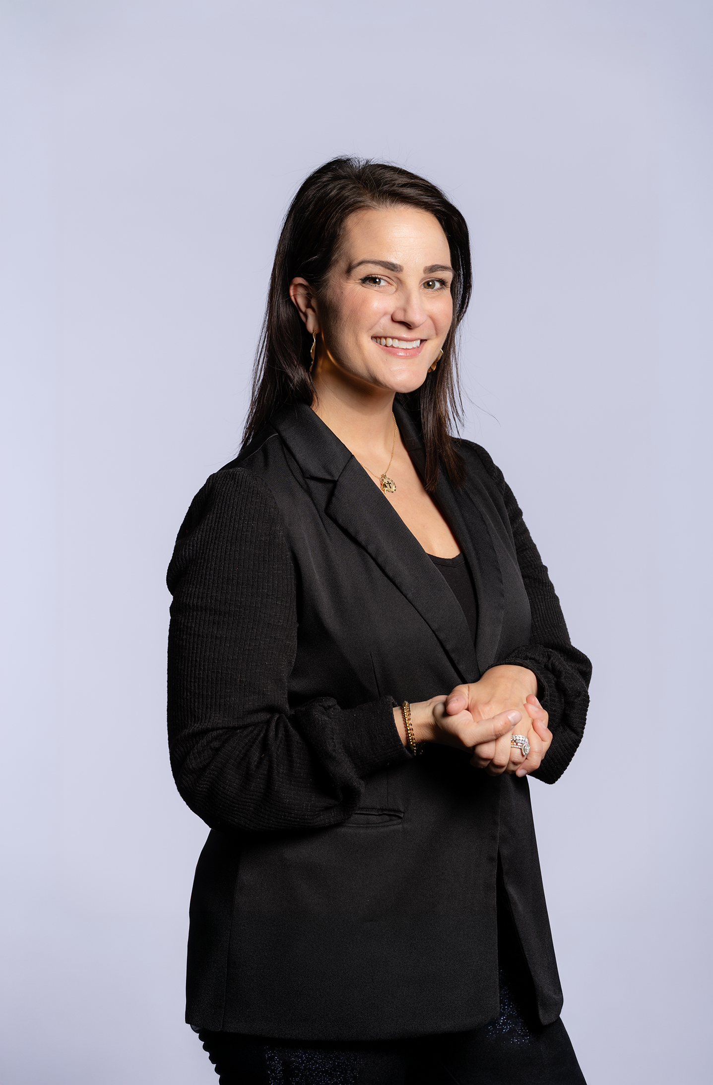 A woman with dark hair smiling, wearing a black blazer and jewelry, standing against a plain light background.
