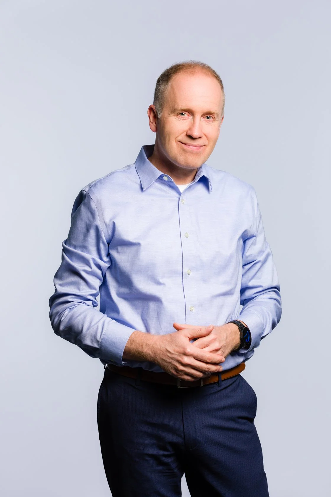 A man in a light blue dress shirt and dark pants standing against a plain light gray background.