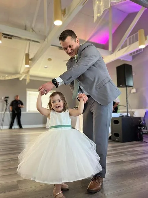 A man in a gray suit dancing with a young girl in a white dress at a celebration or wedding reception in a decorated hall.