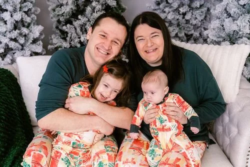Family of four, two adults and two young children, sitting on a white couch in front of decorated Christmas trees, all wearing festive pajamas and smiling.