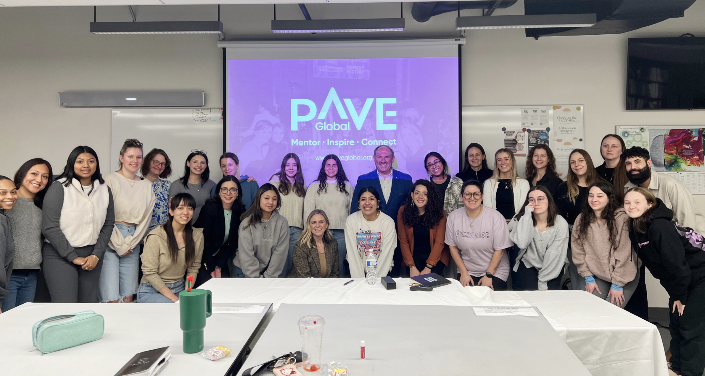 A group of diverse women and men smiling and posing for a photo in a classroom or conference room setting with a large screen displaying 'DAVE Global' and the words 'Mentor, Inspire, Connect' in the background.