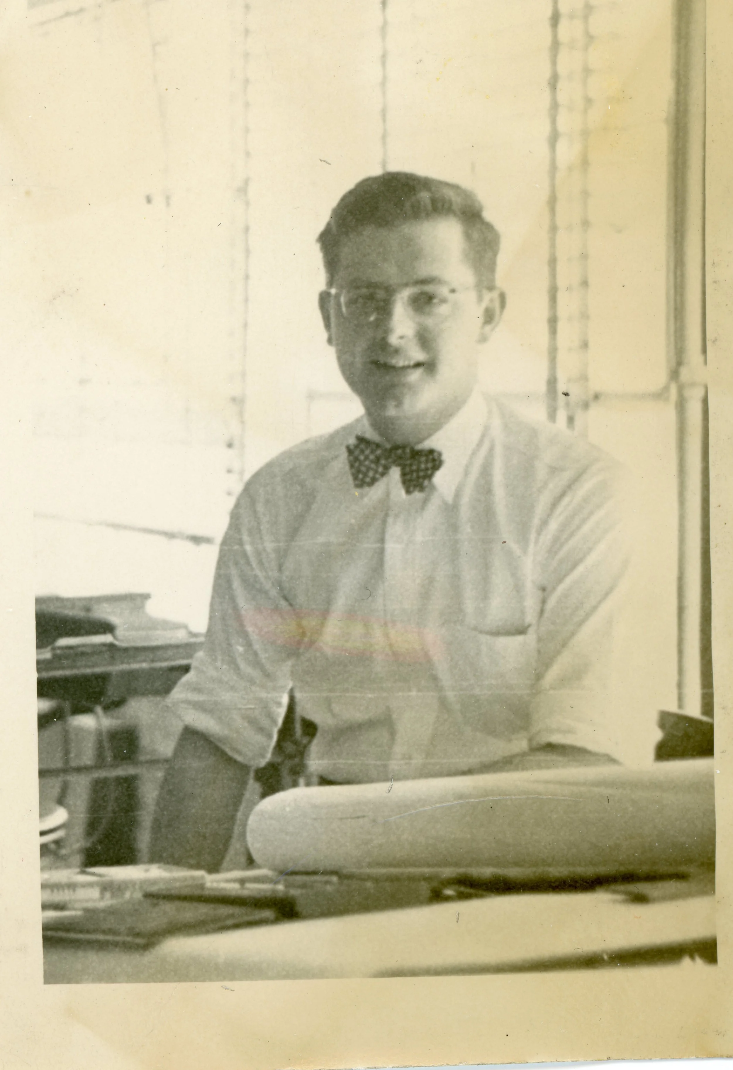 A black and white photo of a young man wearing glasses, a collared shirt, and a polka-dot bow tie, sitting at a desk with papers and office supplies, smiling at the camera.