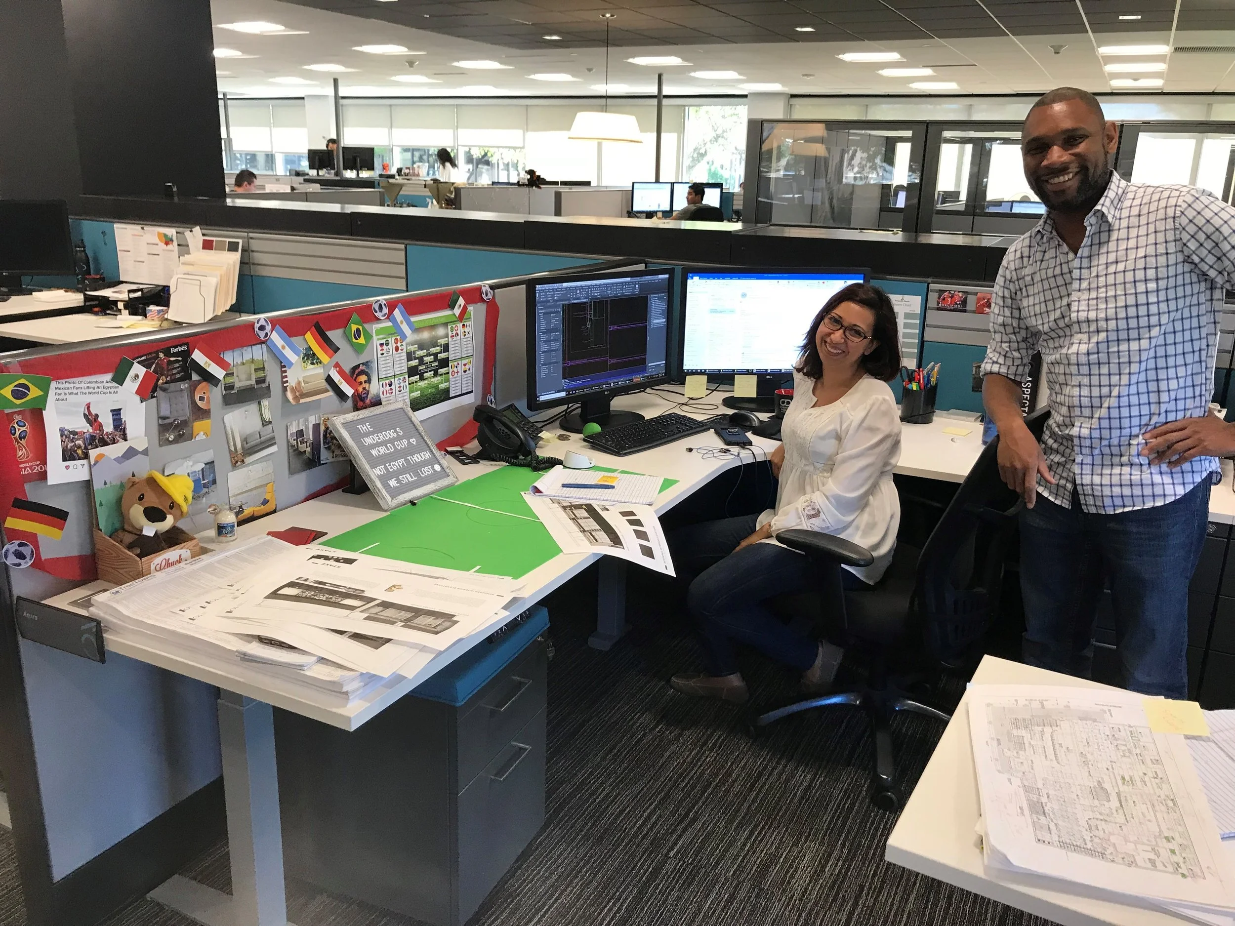 An office cubicle with two women sitting at desks working on computers and a man standing nearby smiling. The cubicle has multiple monitors, papers, and a small display board with flags and pictures.