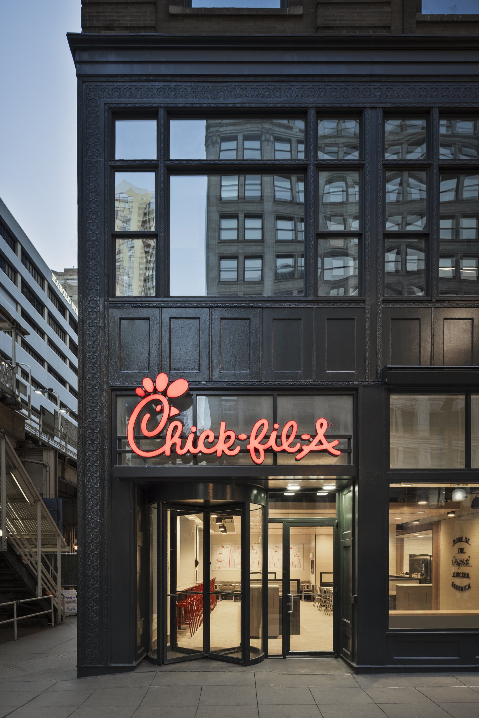 The exterior of a Chick-fil-A restaurant with large glass windows and a black facade. The Chick-fil-A logo with red neon lettering is visible on the front. Inside, red chairs are arranged near the entrance.
