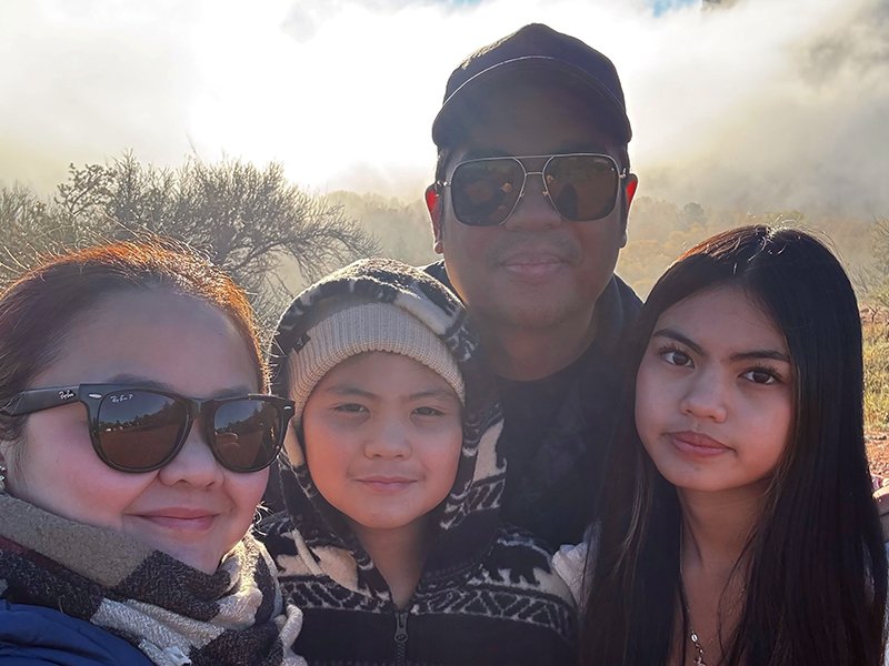 Four people posing outdoors during daytime, with trees and a hazy sky in the background, wearing warm clothing.