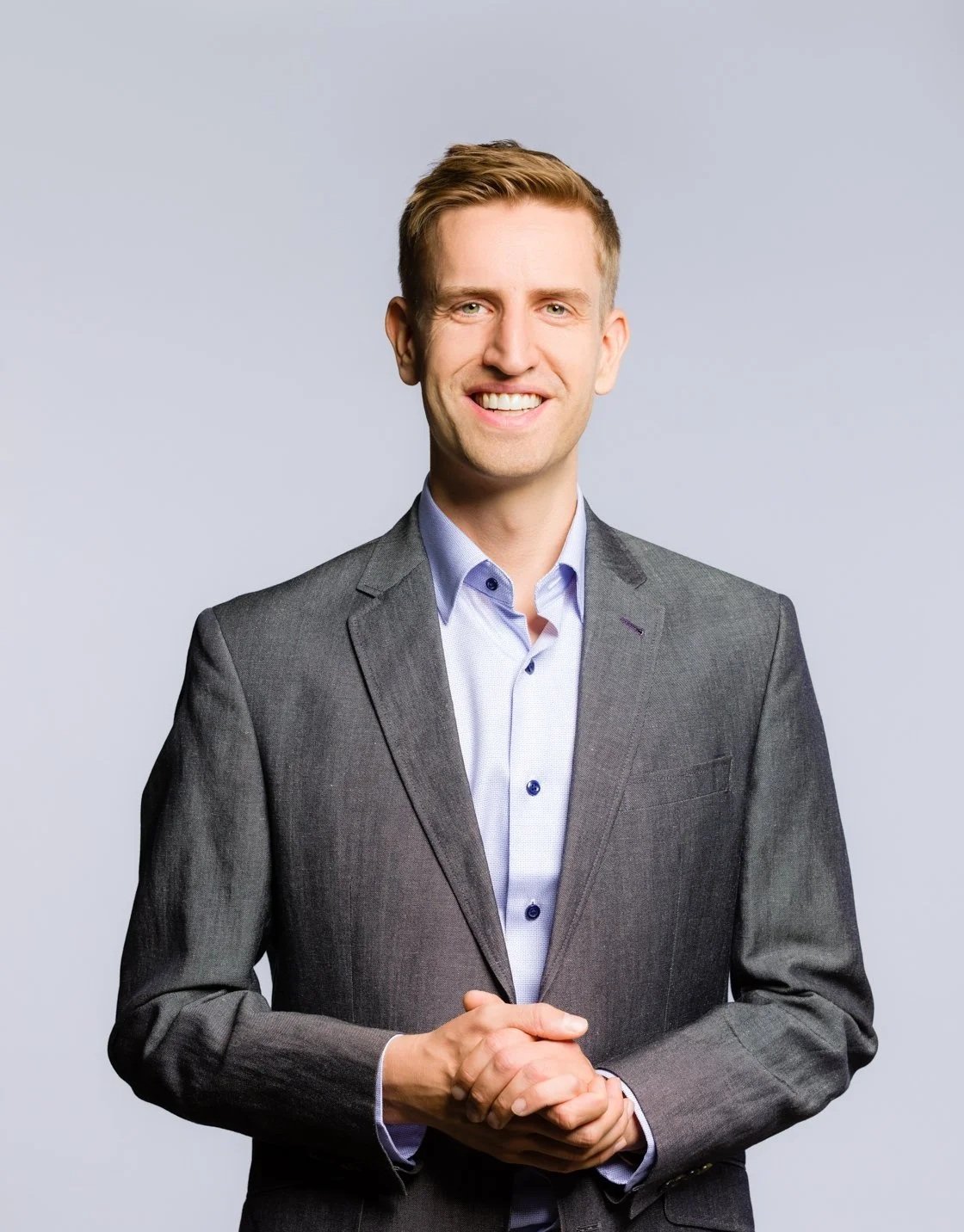 A young man with short blond hair, wearing a dark gray suit jacket and a light blue dress shirt, standing against a plain light gray background, smiling with his hands clasped in front of him.