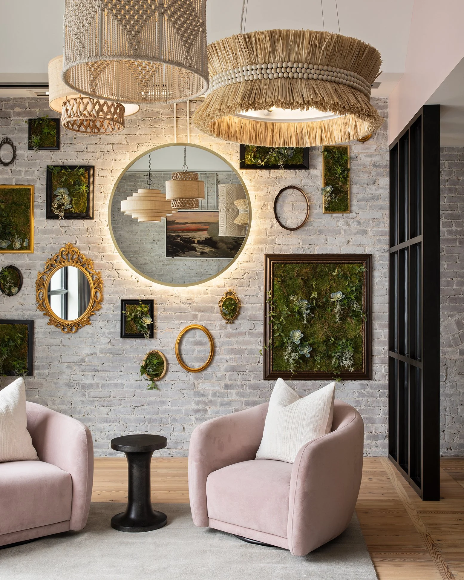 Living room with pink armchairs, black round side table, a large round mirror, and wall art, with hanging woven lampshades and a brick wall decorated with framed green plants and a window.