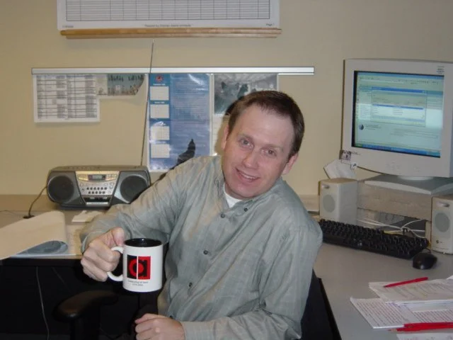 A man sitting at an office desk holding a mug with a red and black logo.
