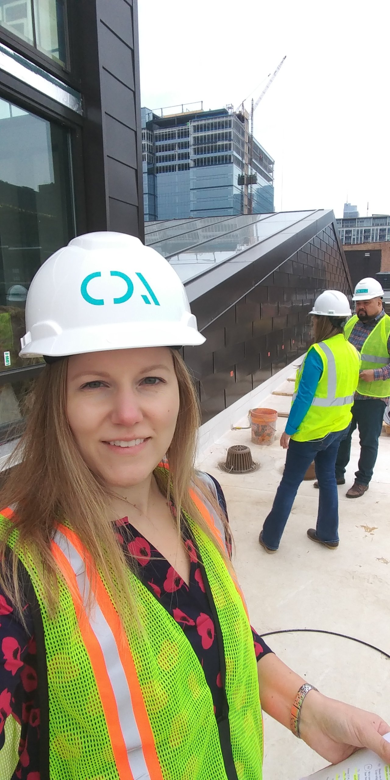 A woman wearing a yellow safety vest and white hard hat smiling at a construction site with other workers in safety gear, a building under construction, and city buildings in the background.