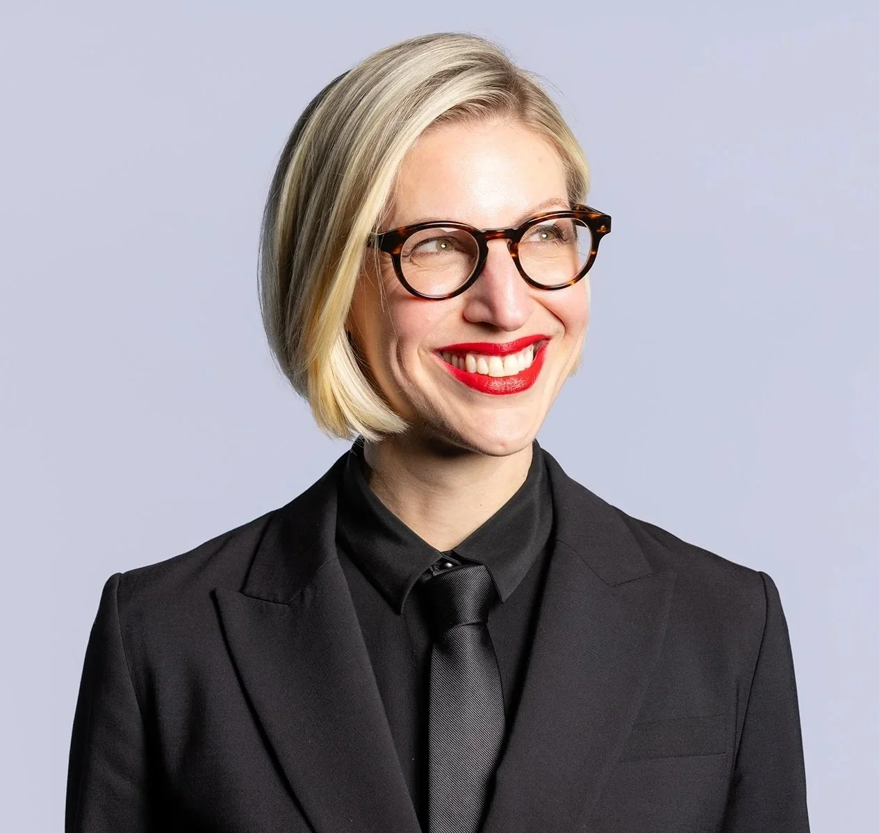 A smiling woman with short blonde hair, glasses, red lipstick, wearing a black suit and tie, against a light gray background.