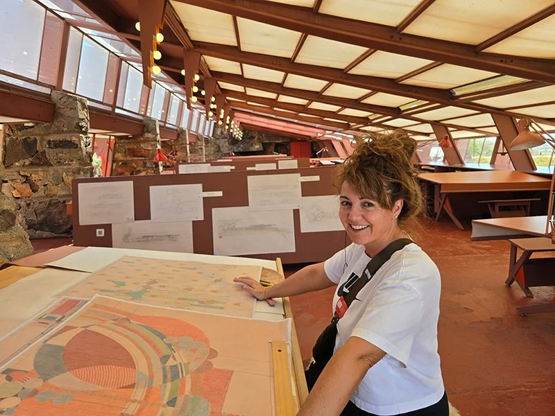 A woman sits at a table with architectural or design drawings, smiling at the camera, in a room with sloped windows and a stone wall.
