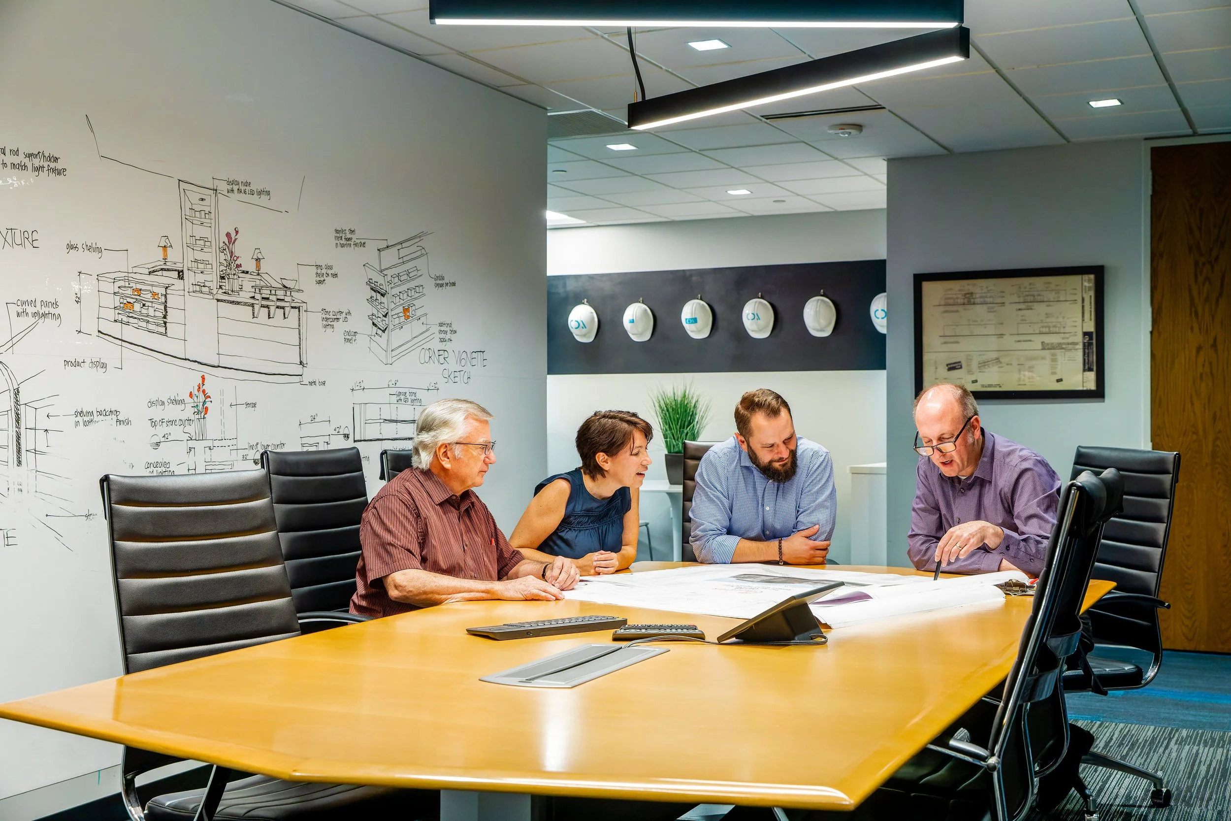 Four people sitting around a conference table in an office, reviewing documents together. A whiteboard with sketches and notes, wall-mounted helmets, and a framed document are in the background.