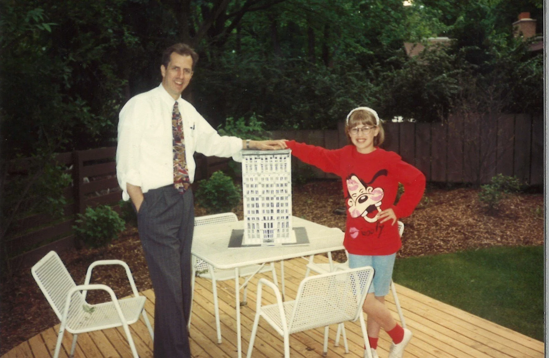 A man and a girl outdoors near a table with a model building. The man is dressed in a white shirt and colorful tie, while the girl is wearing a red sweater with a cartoon face, glasses, and shorts. The girl is smiling and has her arm extended, touching the model building. There are trees and a wooden fence in the background.