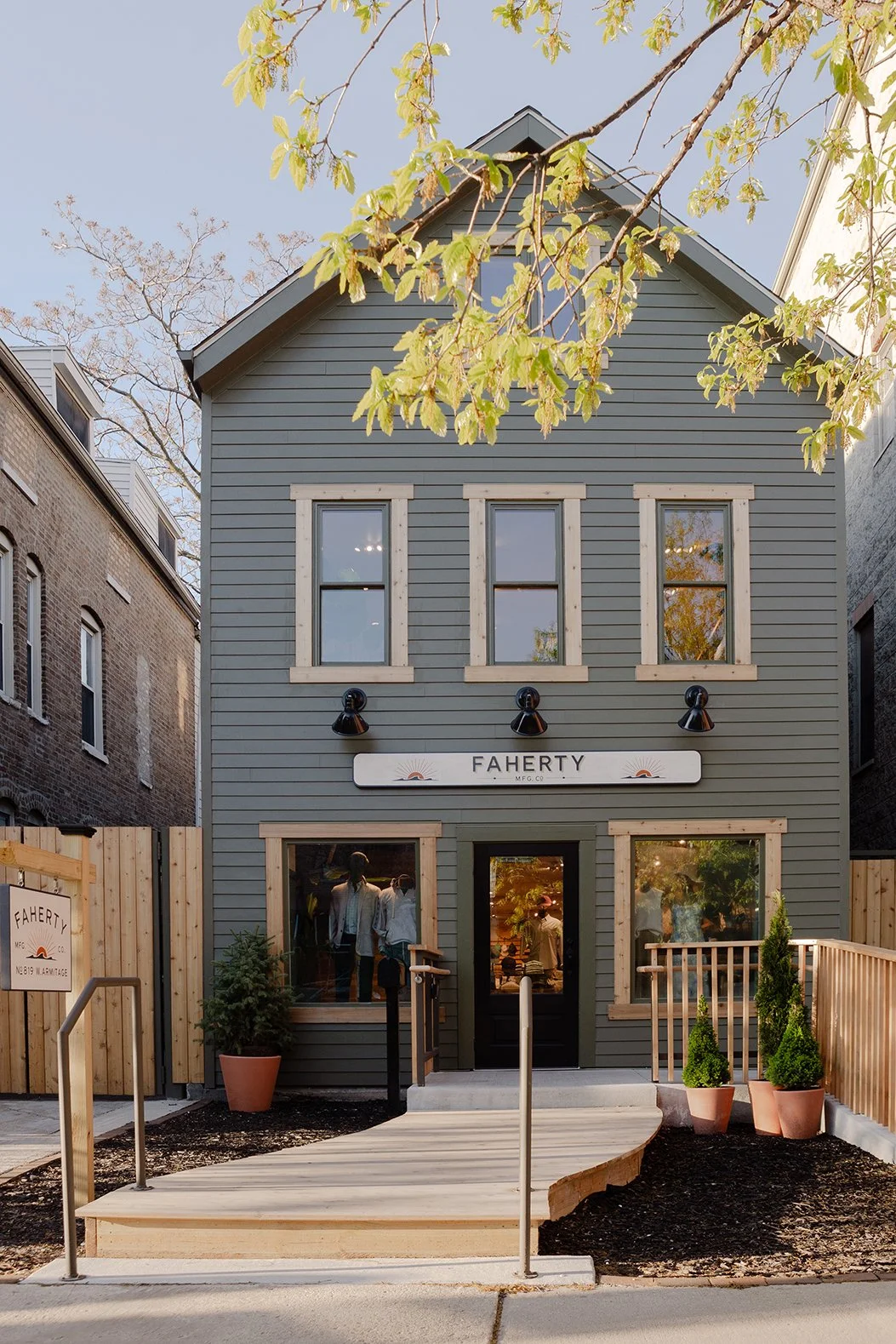 A two-story gray building with beige trim and a sign reading 'FAHERTY' above the entrance, featuring six windows with three on each floor, surrounded by a small front porch with potted plants and a wooden ramp, located between two brick buildings, with a wooden fence on either side and trees overhead.
