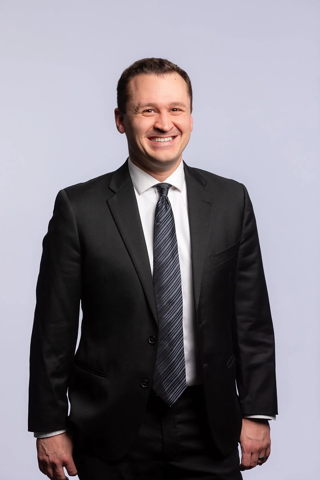 Man in a black suit, white shirt, and patterned tie, smiling against a plain light gray background.