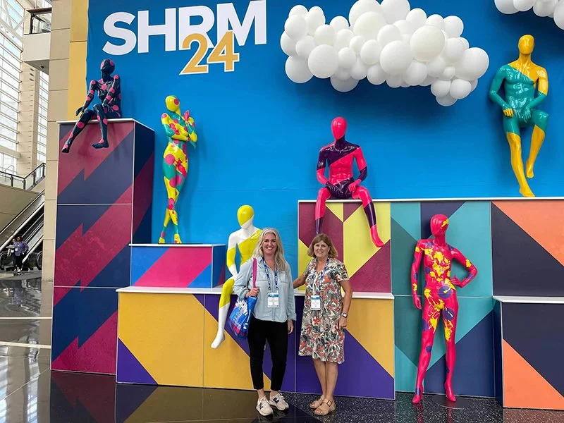 Two women standing in front of colorful mannequin displays at the SHRM 24 conference, with mannequins in bright, patterned suits posed on multicolored platforms.