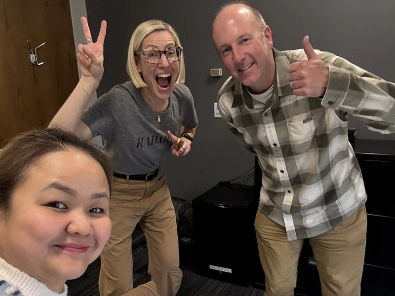 Four people taking a group selfie indoors, smiling and making cheerful gestures.