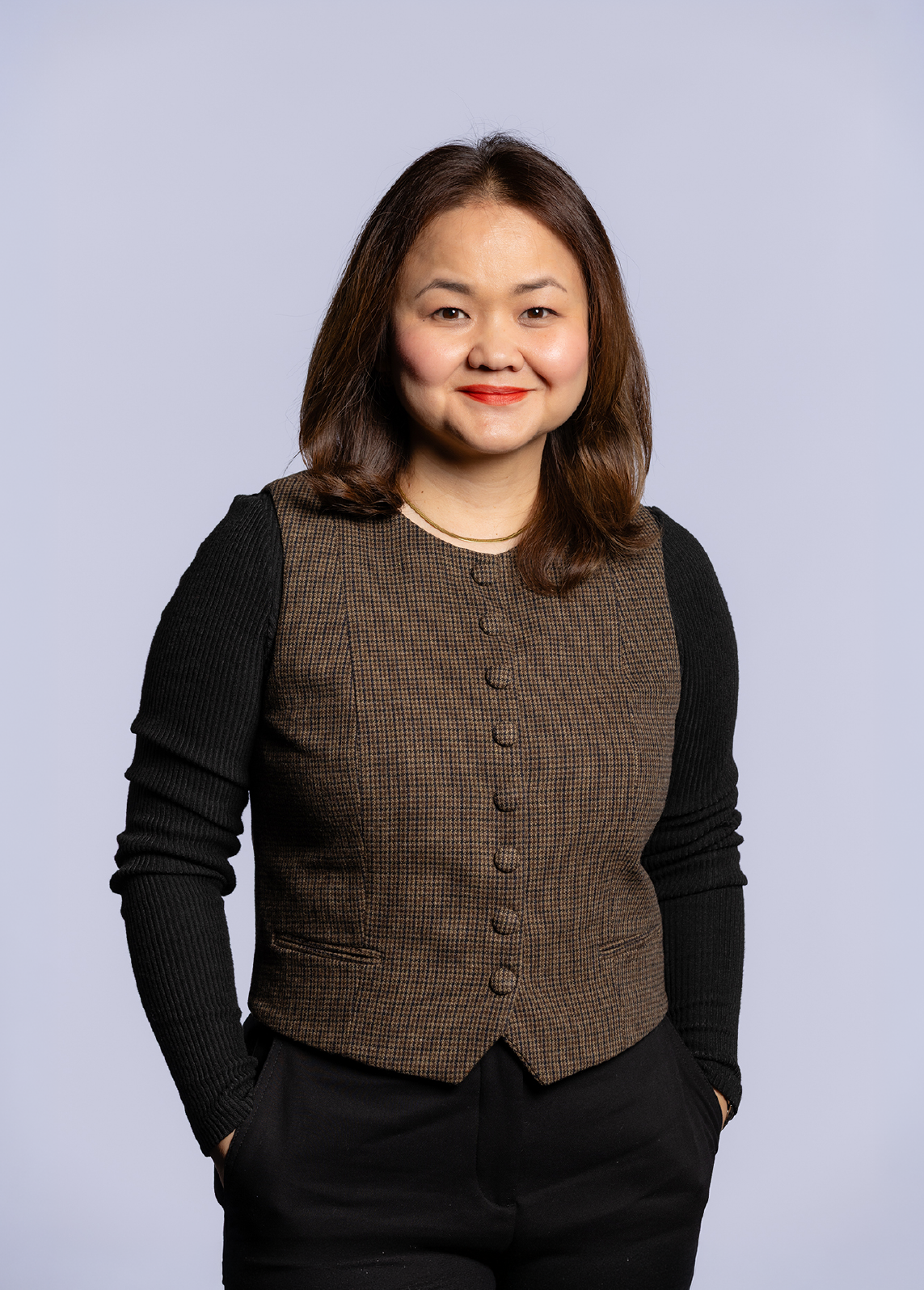 Head and shoulders portrait of an Asian woman with shoulder-length brown hair, smiling, wearing a brown checkered vest with black sleeves, standing against a light gray background.