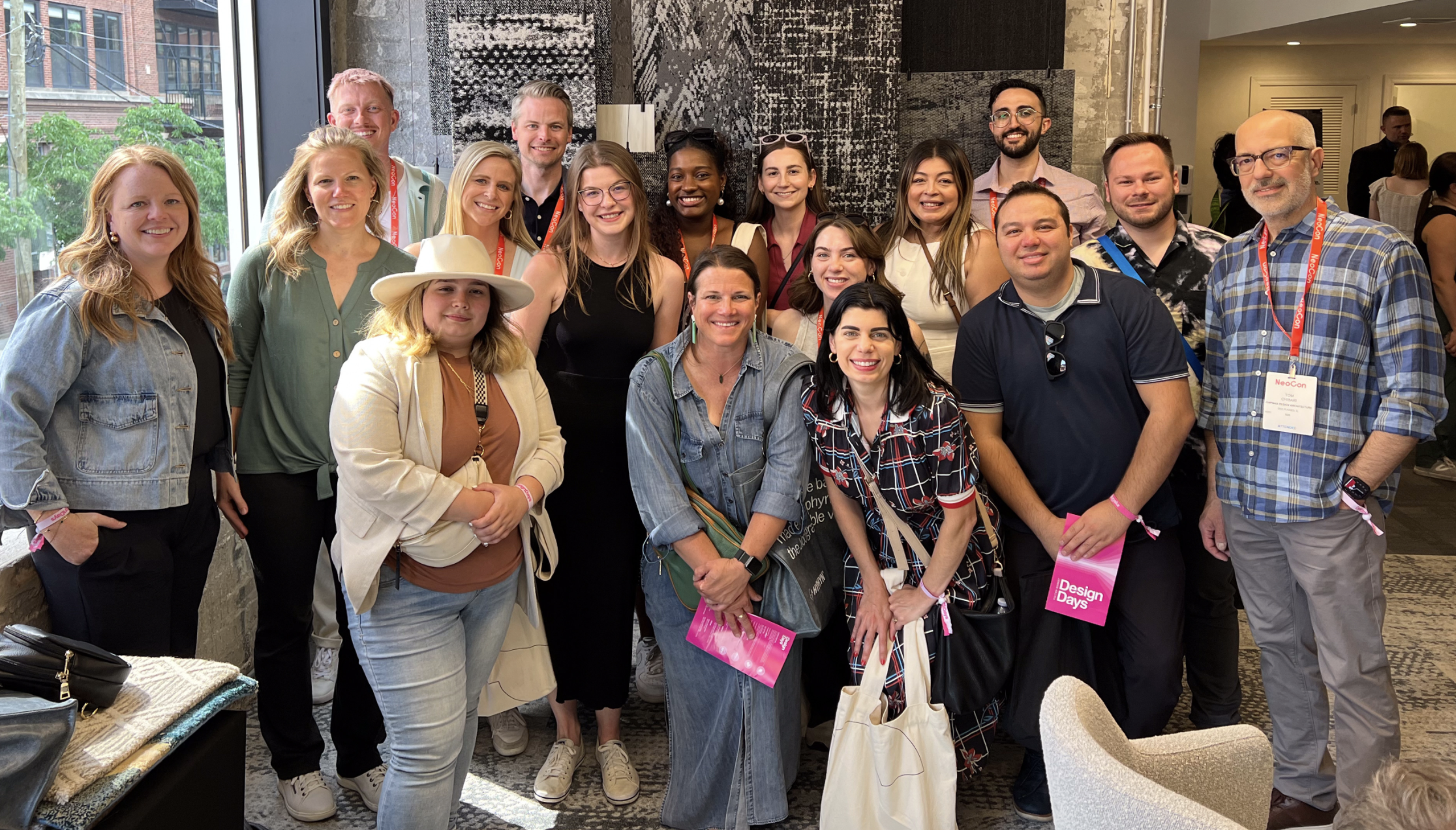 A group of 17 diverse people smiling and posing for a photo in a modern indoor space with large windows, artwork, and a textured black and white wall.