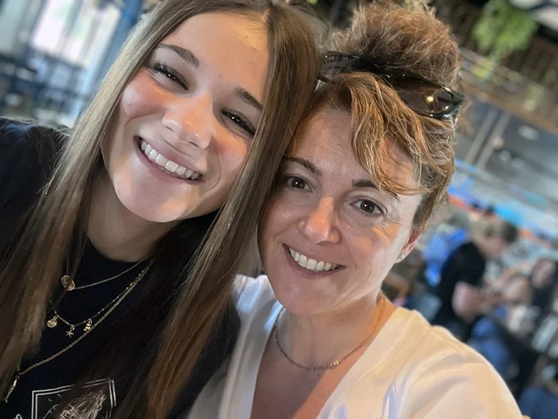 Two women smiling and taking a close-up selfie inside a café or restaurant, with other people in the background.