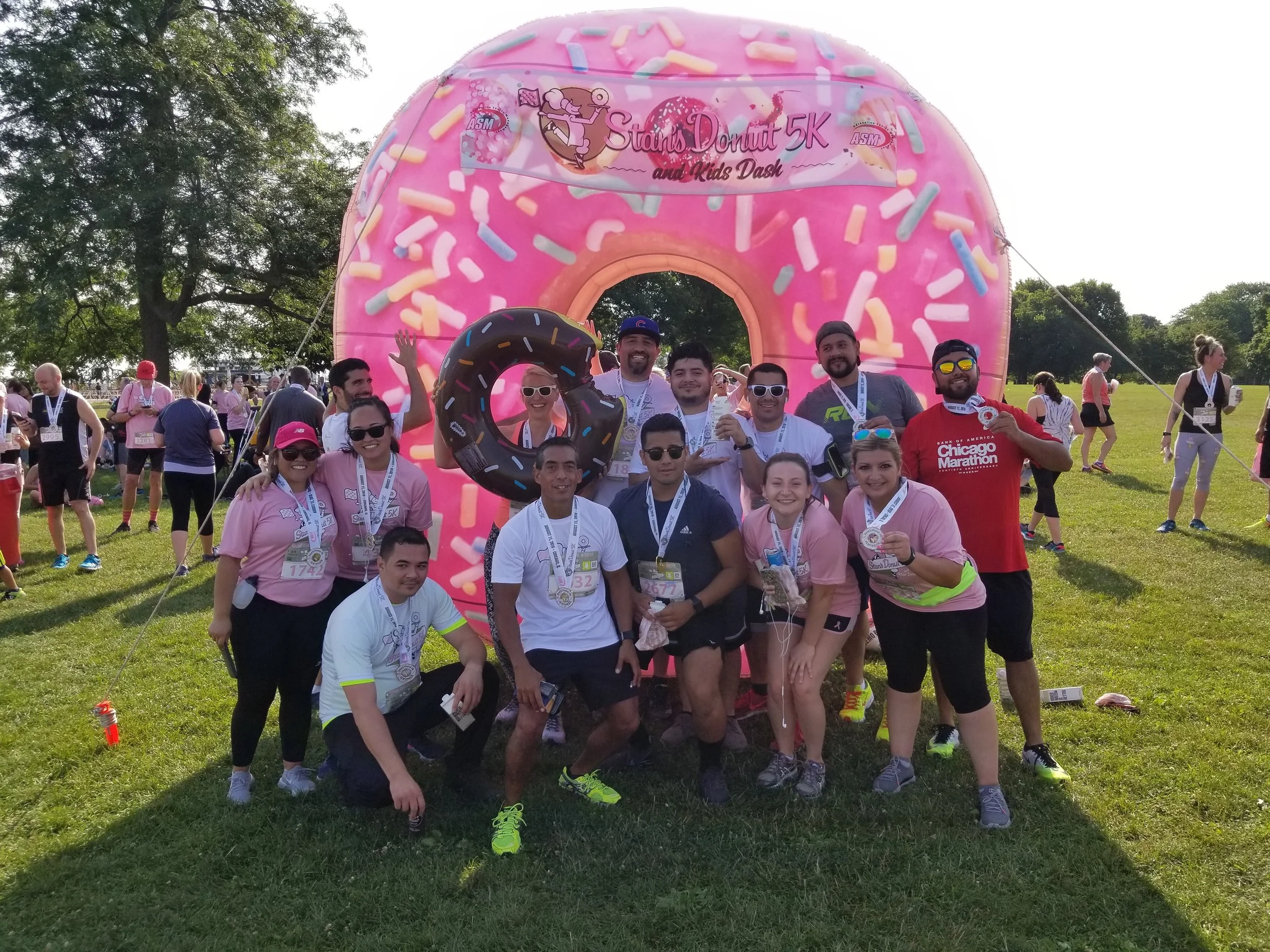 A group of people smiling and posing in front of a large pink donut-shaped inflatable arch during a community running event, with trees and other participants in the background.