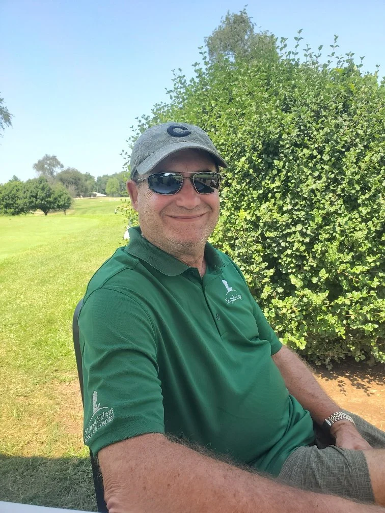 A man wearing sunglasses, a baseball cap, and a green polo shirt sitting outdoors on a sunny day.