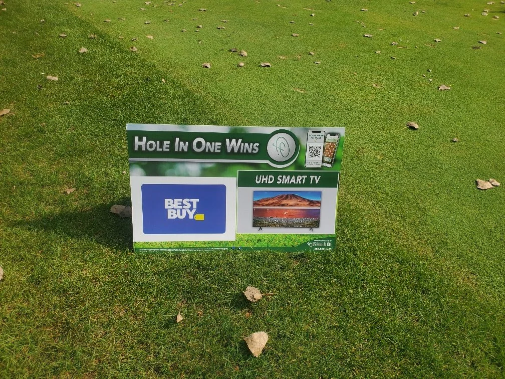 A small sign on a grassy area advertising a promotional golf game called "Hole In One Wins" with a UHD Smart TV and Best Buy logo.