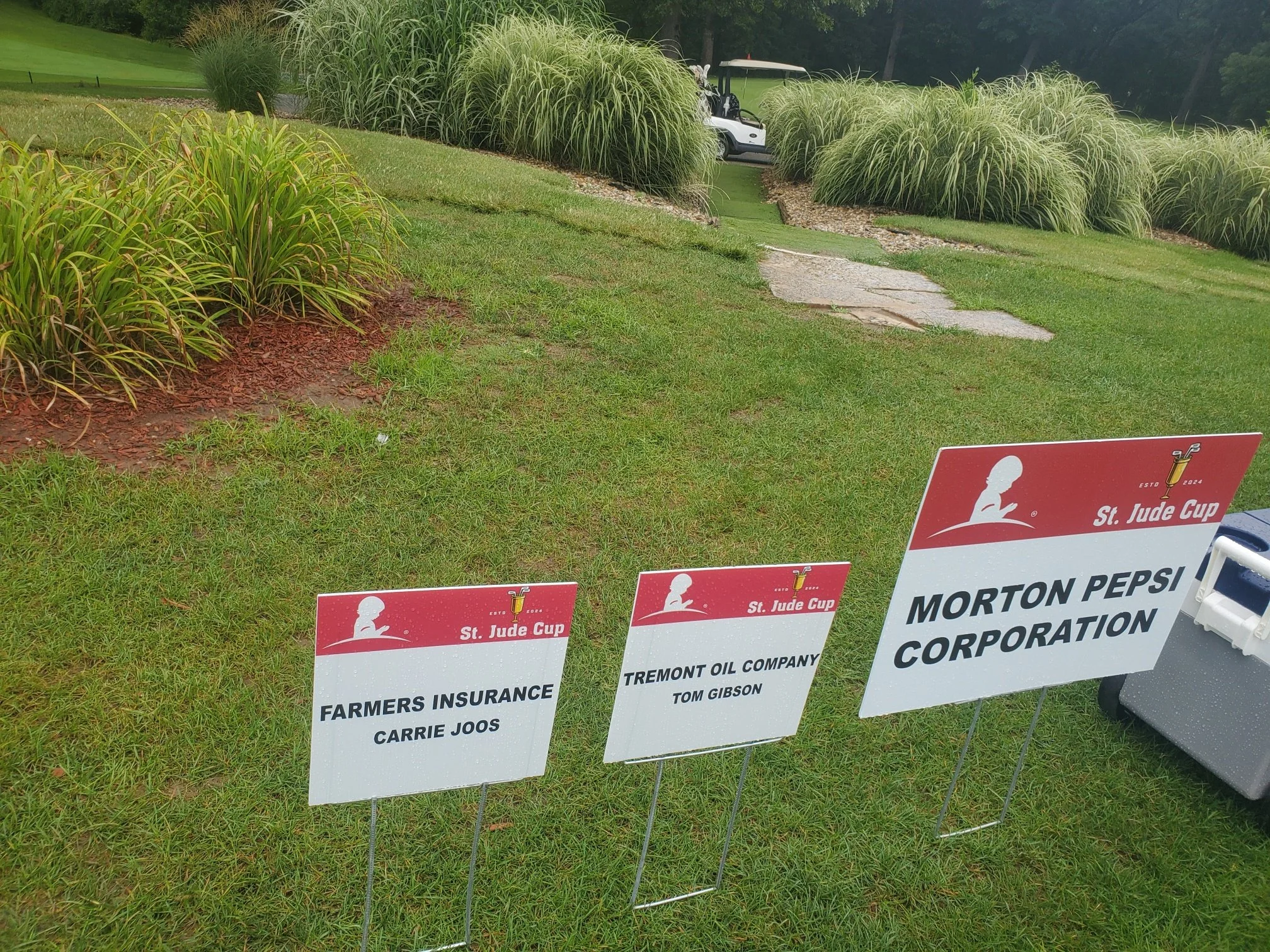 Three sponsor signs on a golf course grass, with a golf cart and stone pathway in the background. The signs are for Farmers Insurance, Tremont Oil Company, and Morton Pepsi Corporation, all affiliated with the St. Jude Cup event.