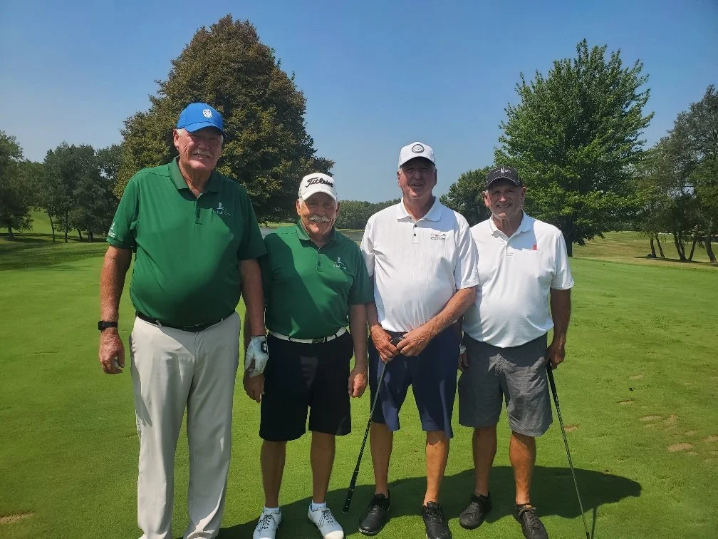 Four men standing on a golf course, posing for a photo. They are wearing golf attire, with three men holding golf clubs. The background features trees and a clear blue sky.