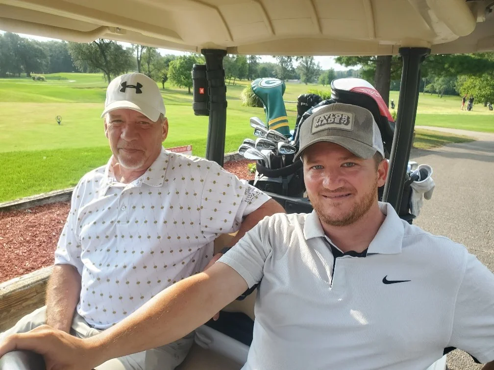Two men sitting in a golf cart at a golf course, surrounded by green grass and trees, with golf clubs in the background.