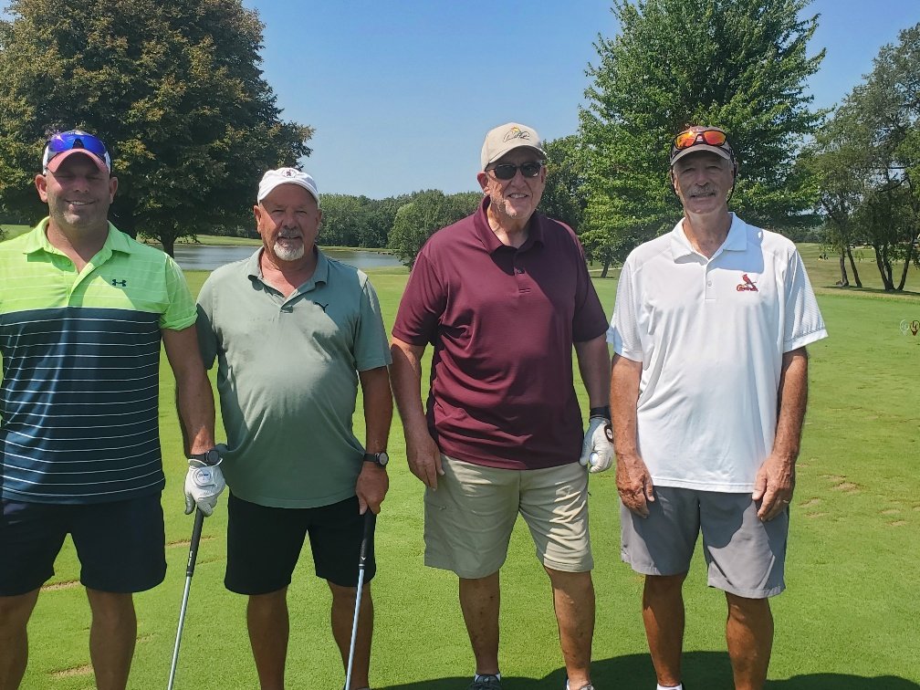 Four men standing on a golf course, holding golf clubs, with trees, grass, and a pond in the background.