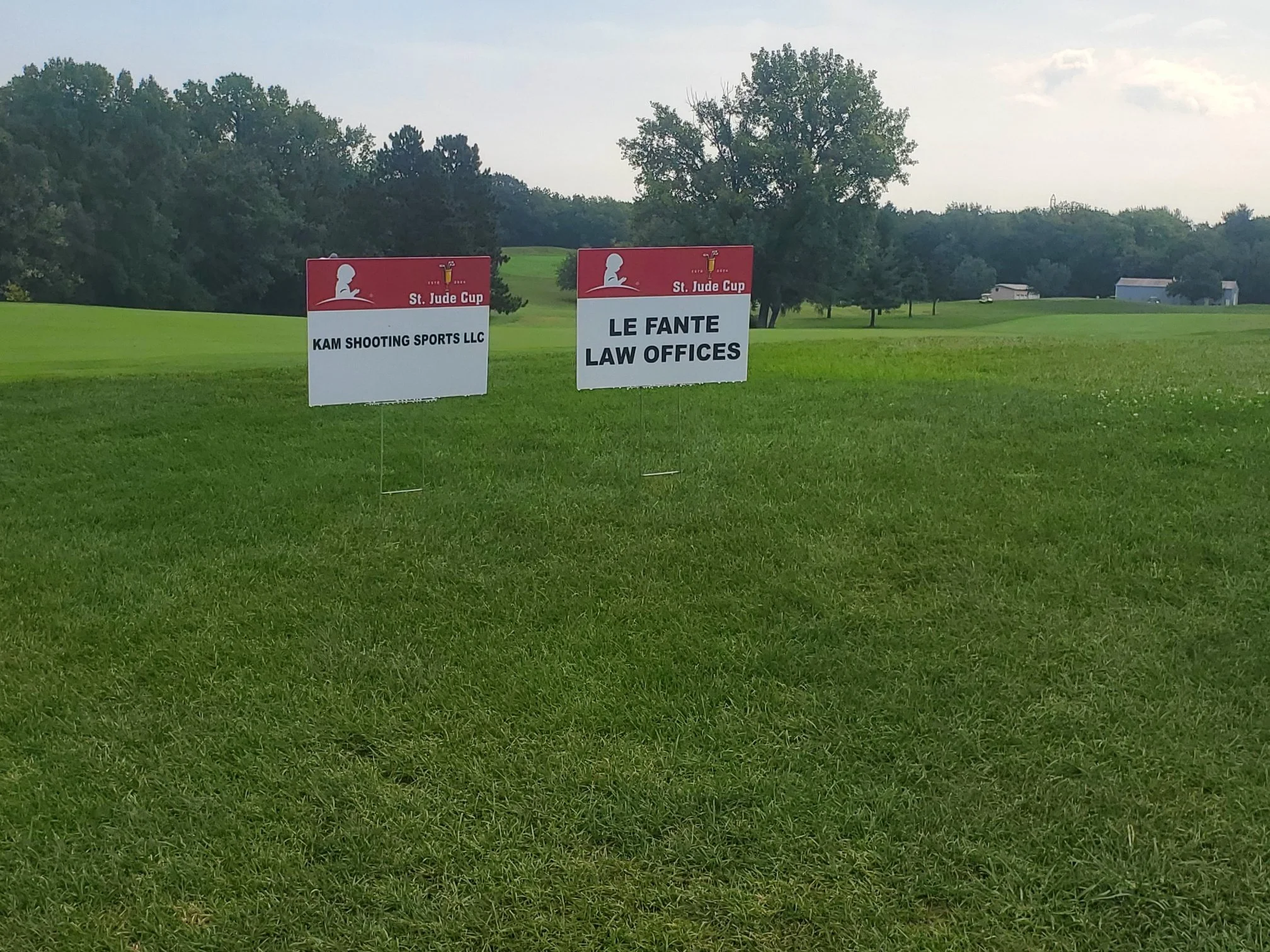 Two signs on a grassy golf course fairway, one for KAM Shooting Sports LLC and the other for Le Fante Law Offices, with trees and a few buildings in the background under a partly cloudy sky.