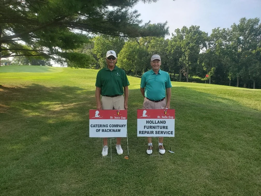 Two men standing on a golf course holding signs for a charity event called St. Jude Cup. One sign reads "Catering Company of Mackinaw" and the other reads "Holland Furniture Repair Service." The men are wearing golf attire and caps, with trees and a 